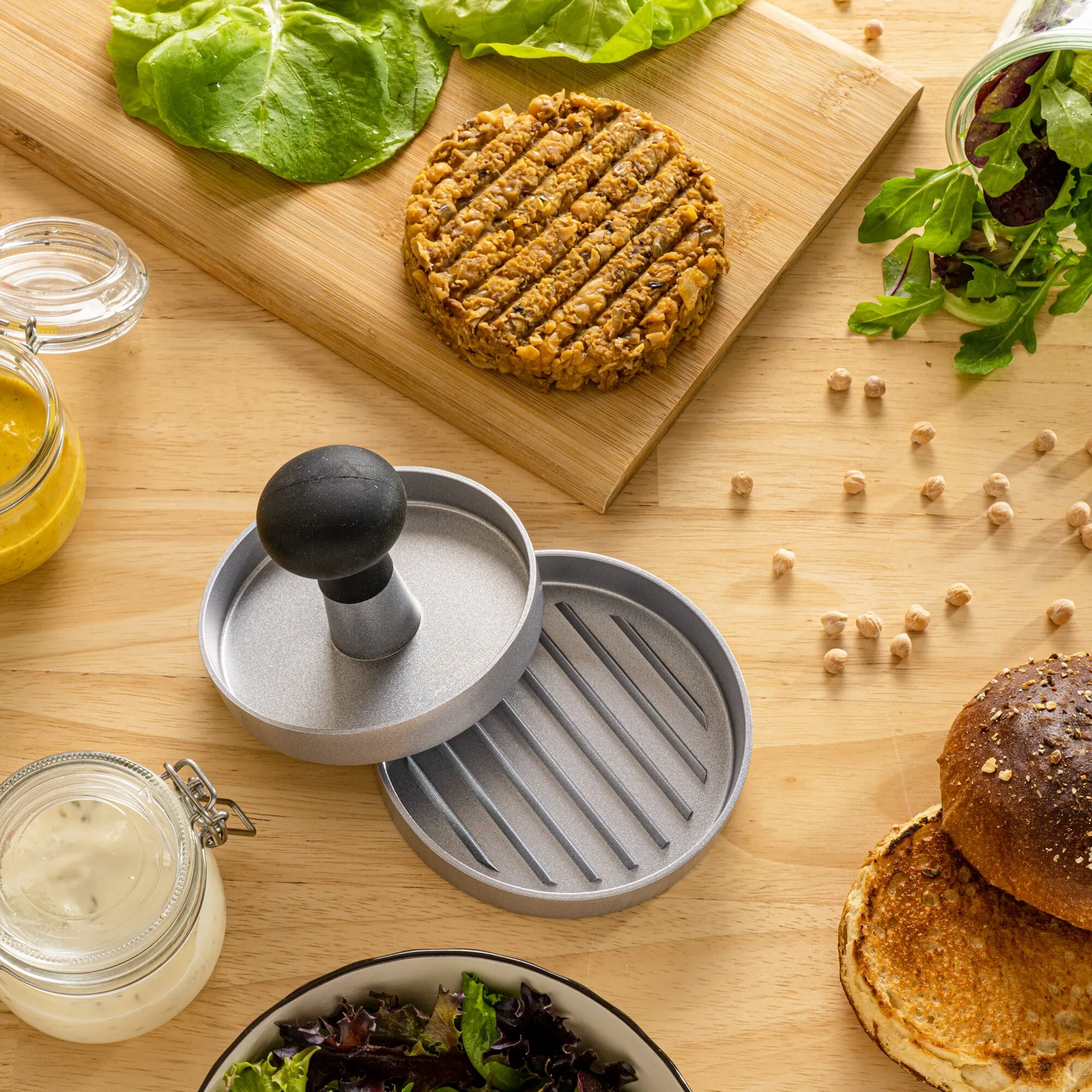 Graue Hamburgerpresse mit geriffelter Bodenplatte und schwarzem Griff neben geformtem Patty, Salat, Brötchen und Saucen auf Holztisch