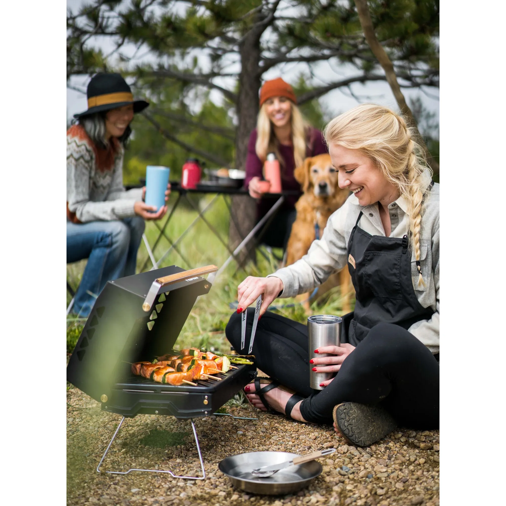 Mobile Gasgrill Kuchoma mit geöffnetem Deckel, auf dem Gemüsespieße gegrillt werden; eine Frau bedient ihn im Freien mit Grillzange