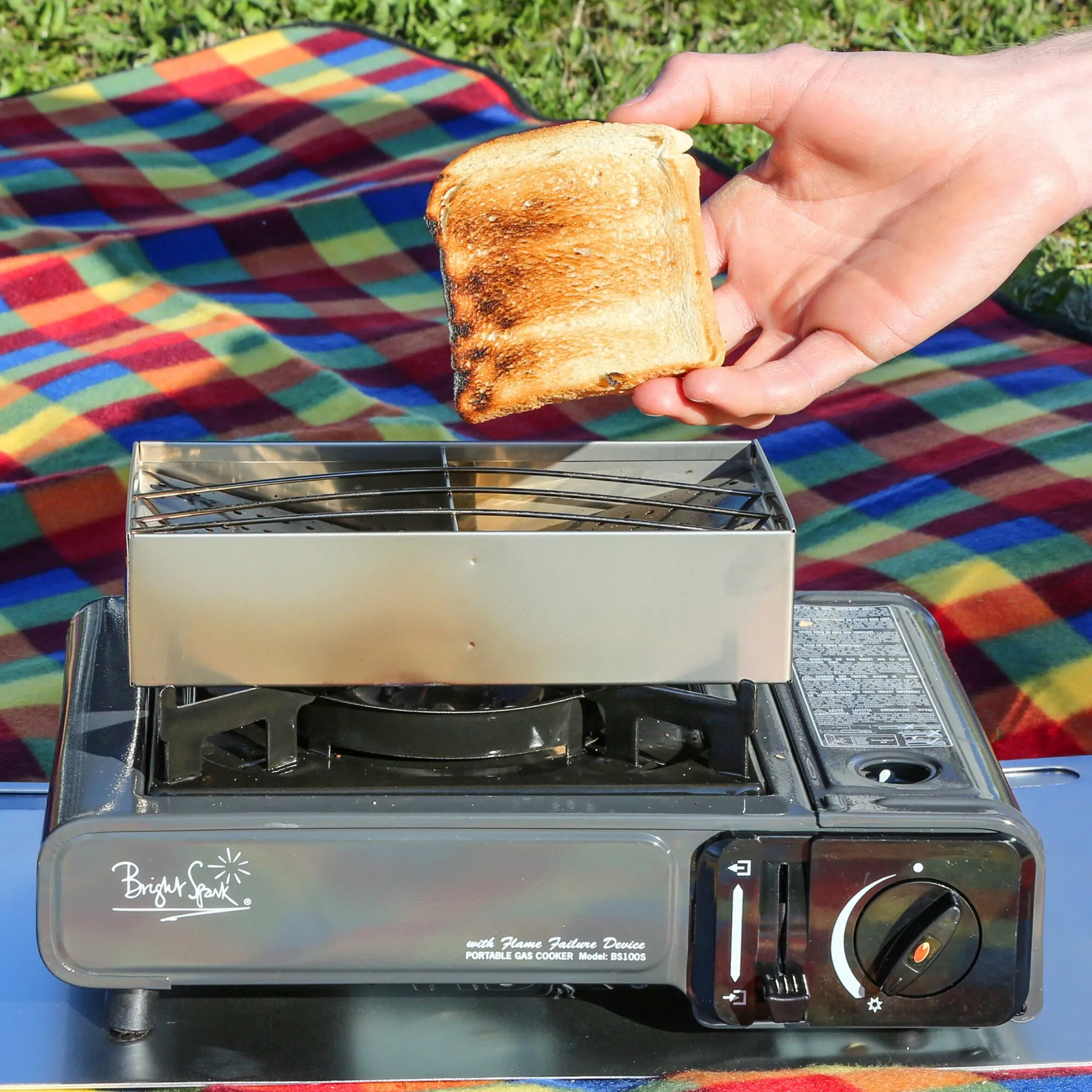 CAMP-A-TOASTER® aus Edelstahl auf einem tragbaren Gaskocher, während eine Hand getoastetes Brot darüber hält