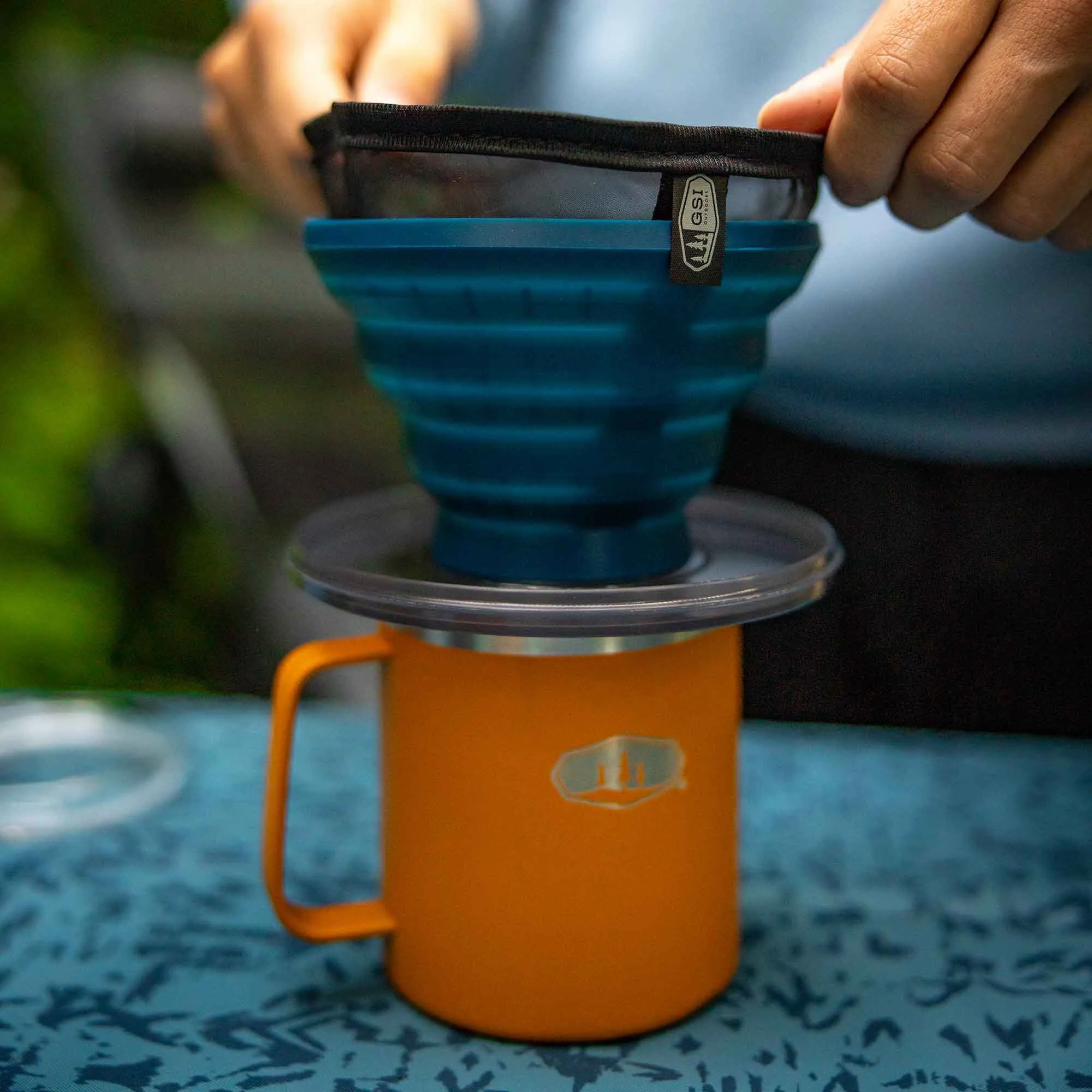 Schwarzer wiederverwendbarer Kaffeefilter in blauem Handfilter über einer orangefarbenen Tasse, von Händen gehalten