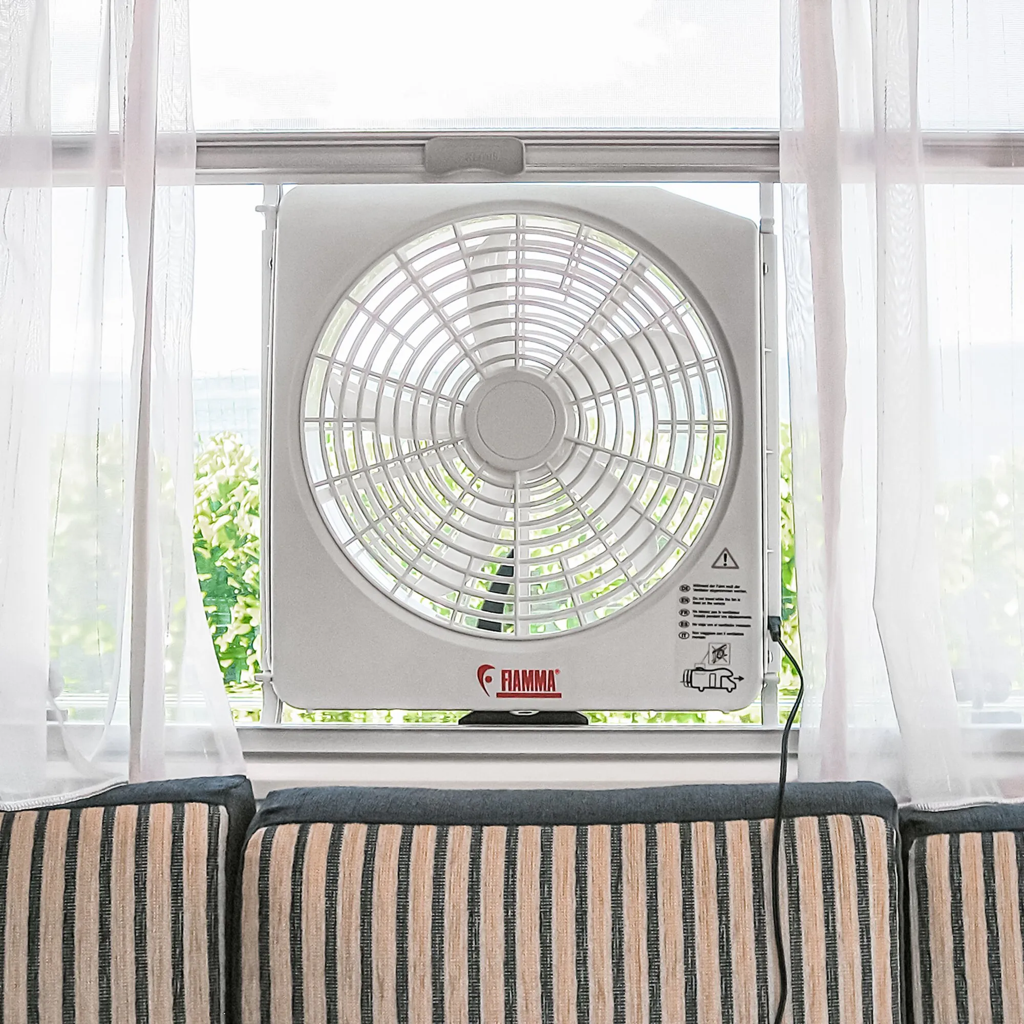 Weißer Fiamma-Dachhaubenventilator Turbo Kit mit Schutzgitter, montiert vor einem Fenster im Wohnmobil vorn an Sitzpolstern