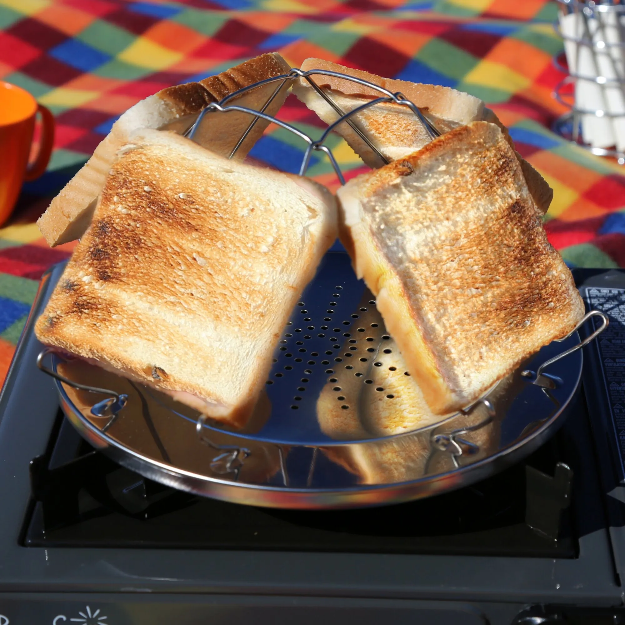Edelstahl-Campingtoaster mit perforierter Grillfläche auf einem Gaskocher, darauf mehrere goldbraun geröstete Brotscheiben