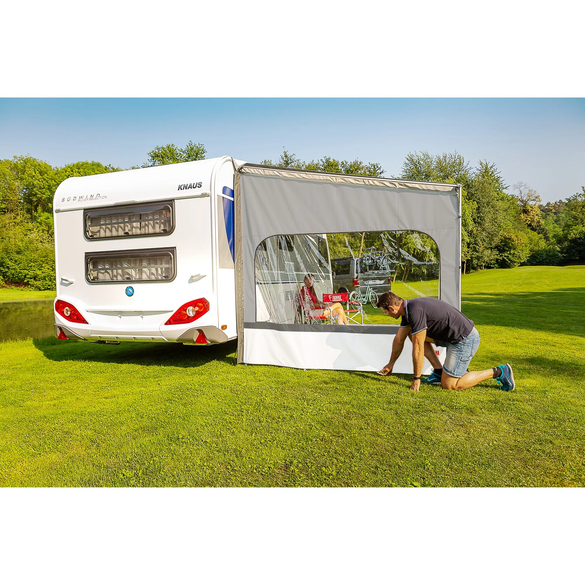 Graue Seitenwand Side W CS, 225 cm, am weißen Knaus-Wohnwagen montiert; ein Mann befestigt sie auf einer Wiese