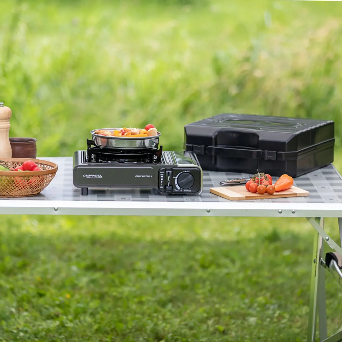Grüner Campingkocher Camp’Bistro 3 mit Pfanne und zubereitetem Gemüse auf einem Campingtisch neben einer schwarzen Transportbox