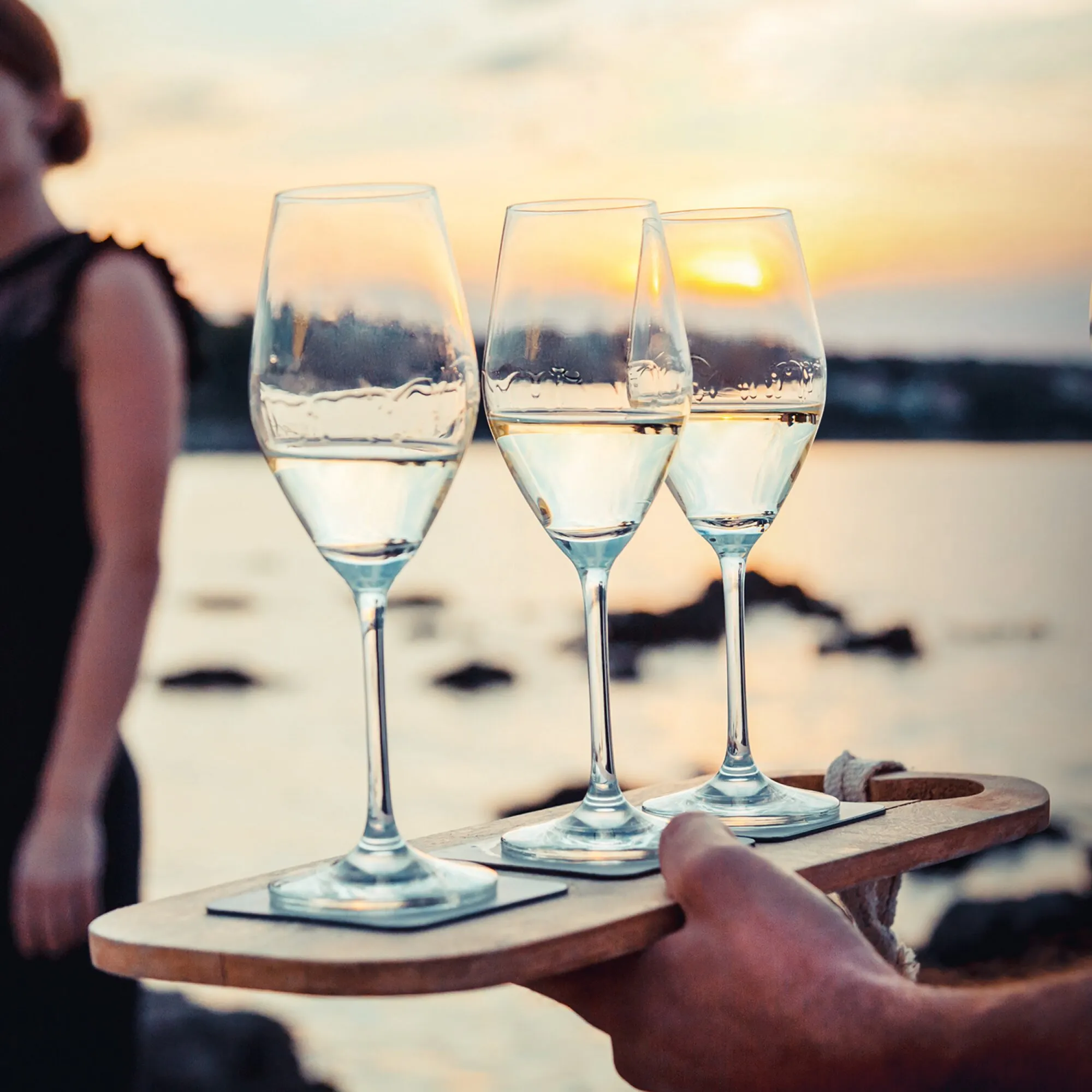 Drei transparente Magnet-Kristallgläser in Champagnerglasform mit hellem Getränk auf einem Holztablett vor dem Sonnenuntergang