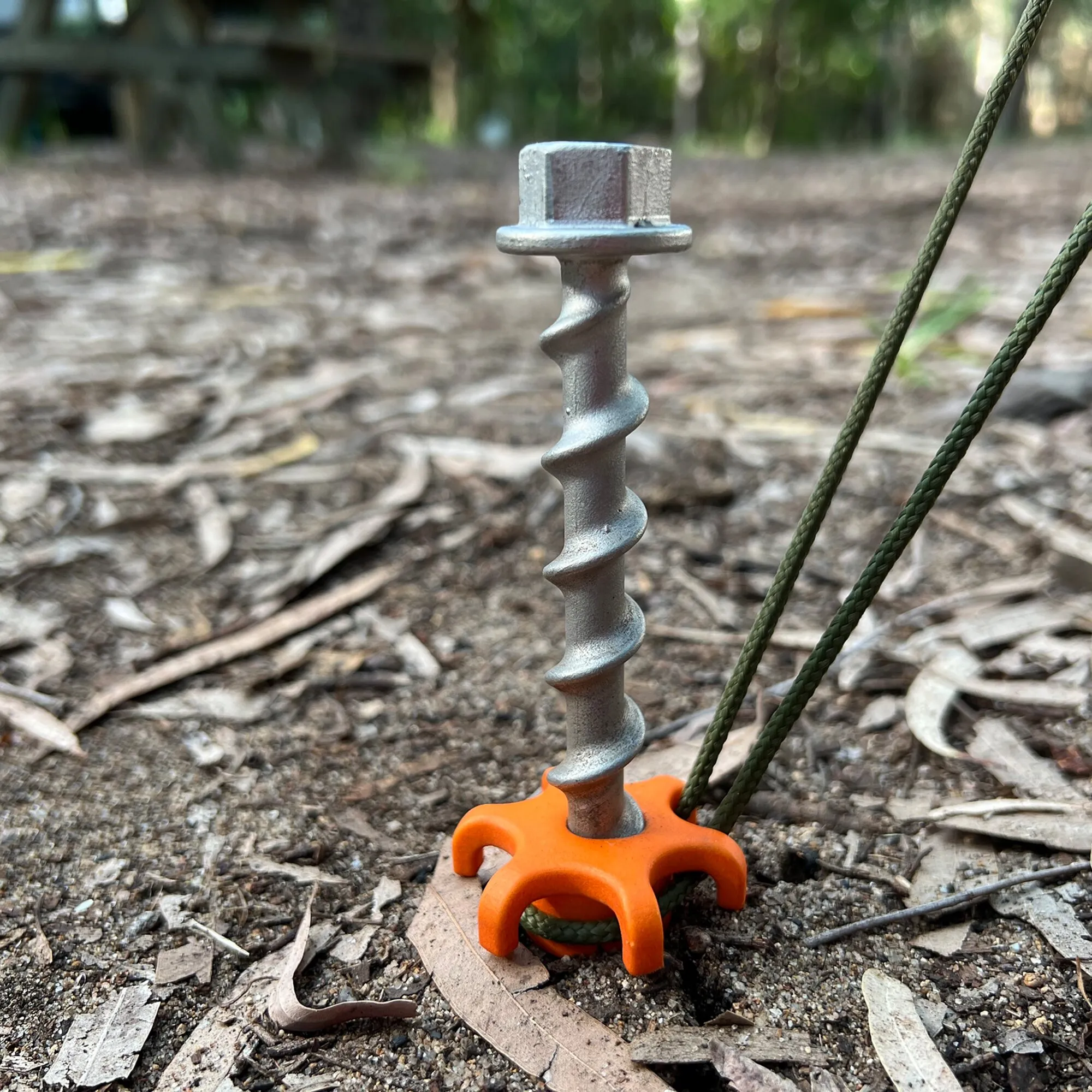 Metallener PP Schraubhering Peg&Stop HeavyDuty mit orangefarbener Kunststoffauflage im Waldboden, daran befestigte grüne Seile