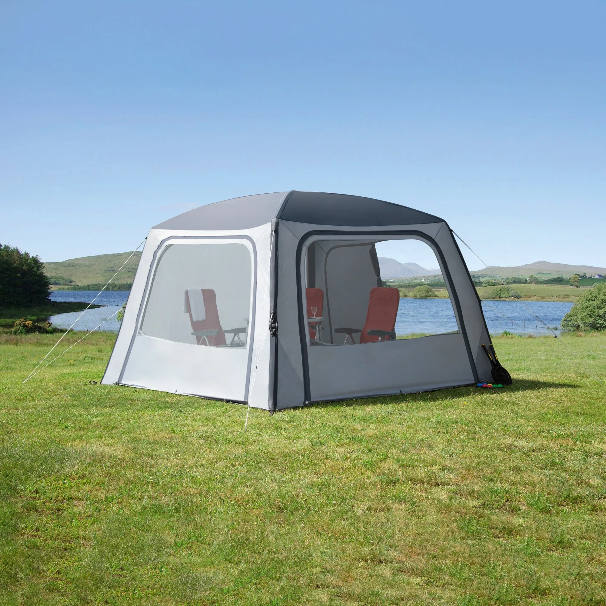 Pavillon Relax Air in Grau und Weiß mit transparenten Fenstern, roten Campingstühlen und Tisch auf einer Wiese am See