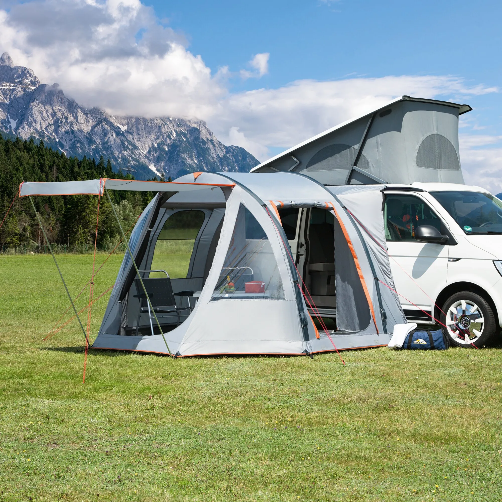 Busvorzelt Picco Air in Grau mit orangefarbenen Akzenten, aufgebaut an einem weißen Campervan auf einer Wiese vor Bergkulisse