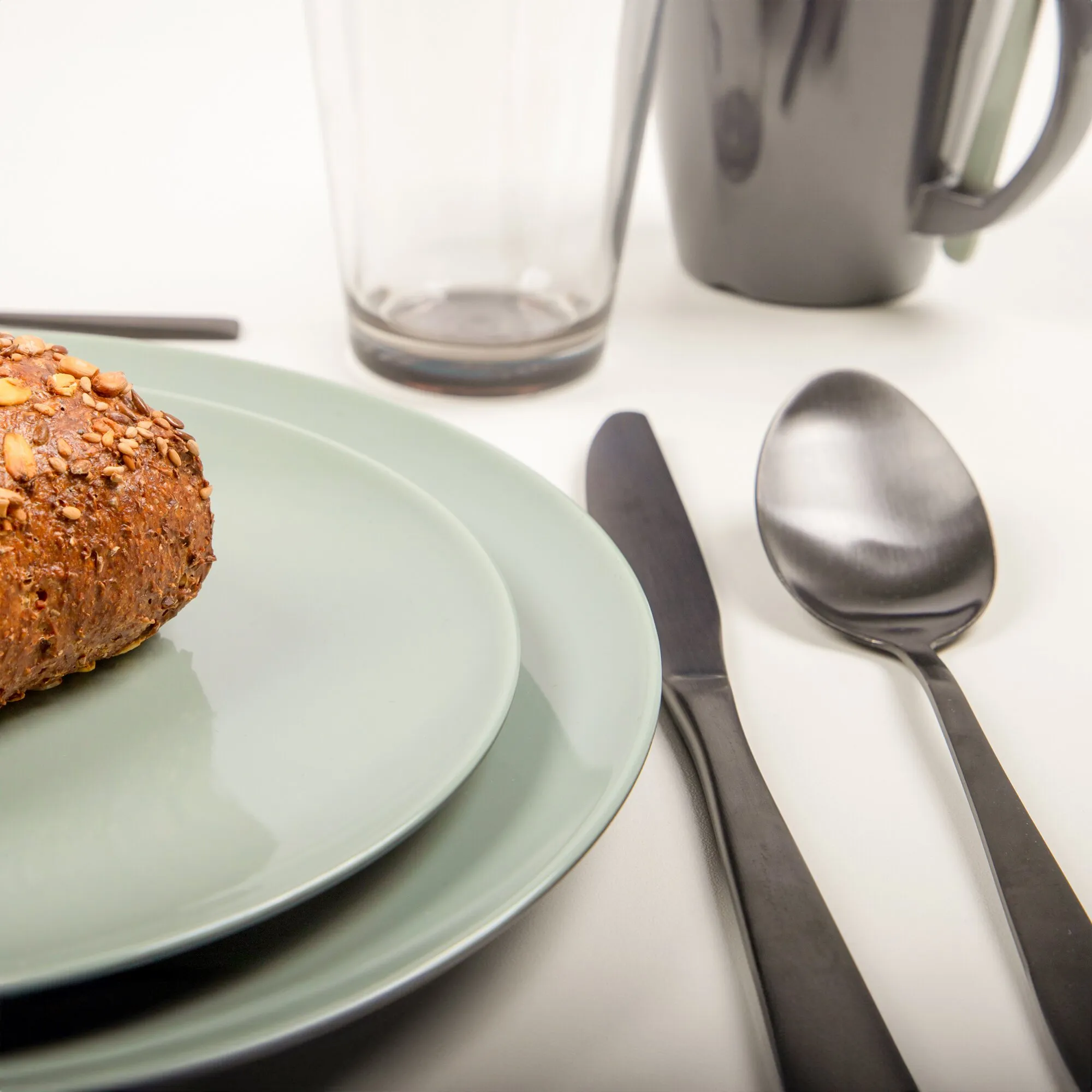 Hellgrünes Geschirrset Vivid Line mit zwei Tellern, einem Brot mit Körnern, Glas, Tasse und schwarzem Besteck gedeckt