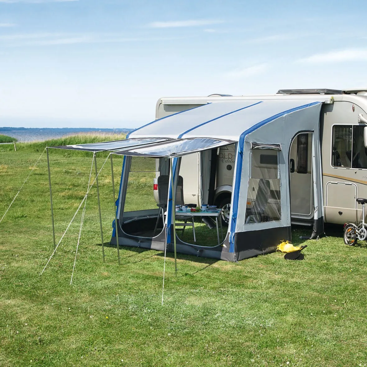 Blau-graues Teilzelt Space Air HQ am Wohnmobil mit geöffneten Seitenwänden, Fenstern, Sonnendach und Campingmöbeln auf einer Wiese am Meer