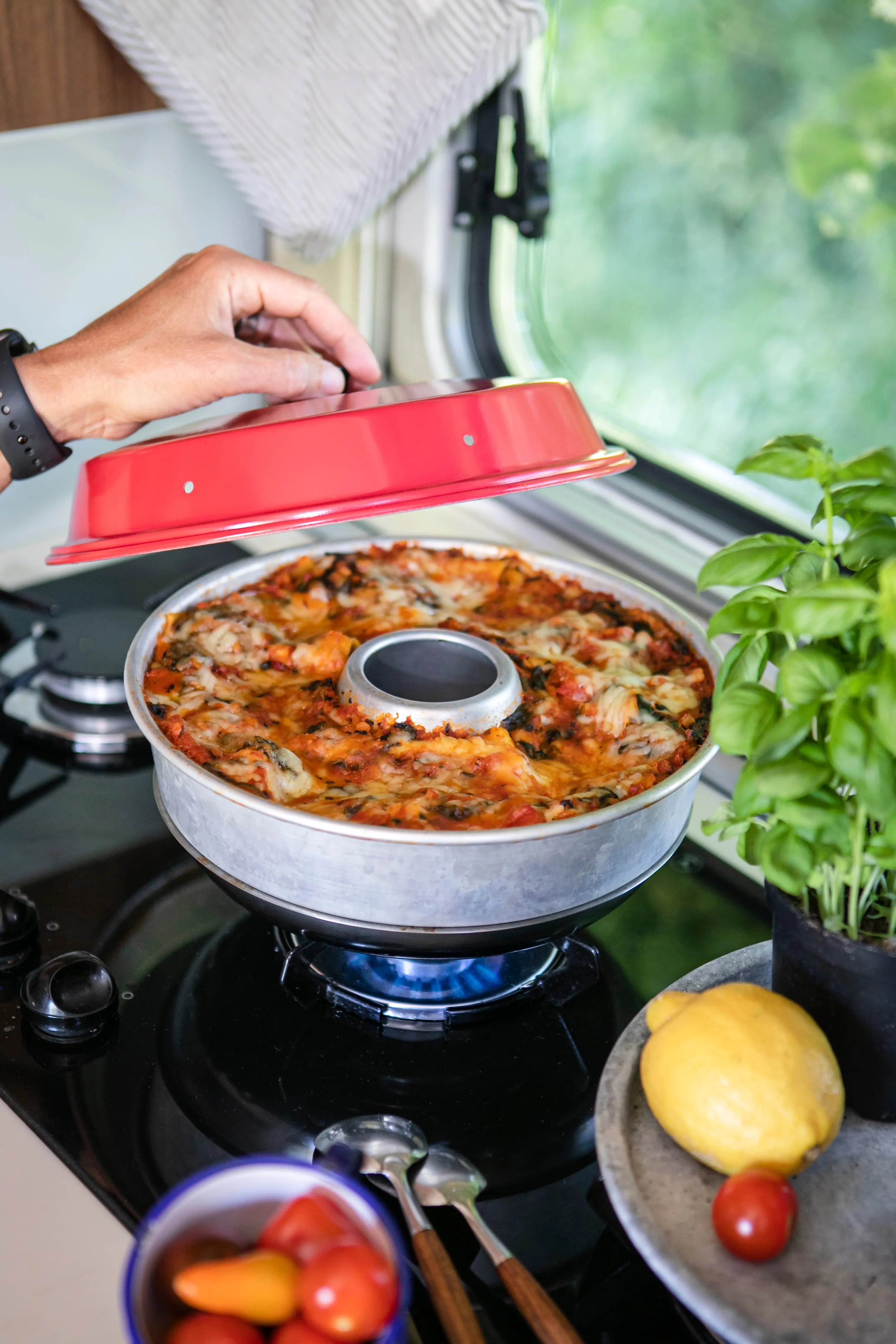 Roter Backofen Classic mit geöffnetem Deckel über einer runden Metallform mit gebackener Speise und Händen am Gerät
