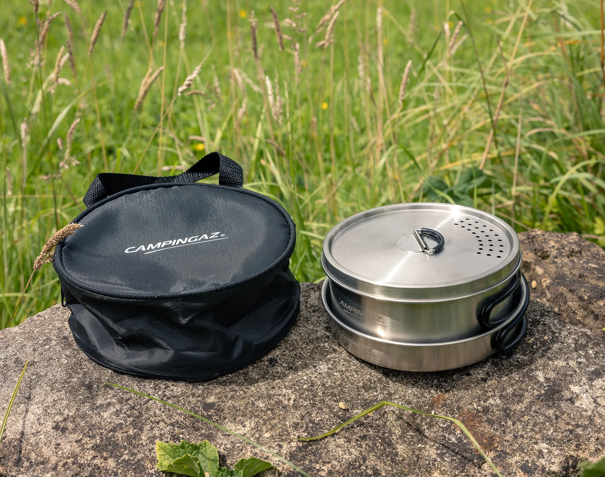 Edelstahl-Trekking-Kochgeschirrset mit Deckel und Griffen neben schwarzer CampinGaz-Tragetasche auf einem Felsen im Grasland