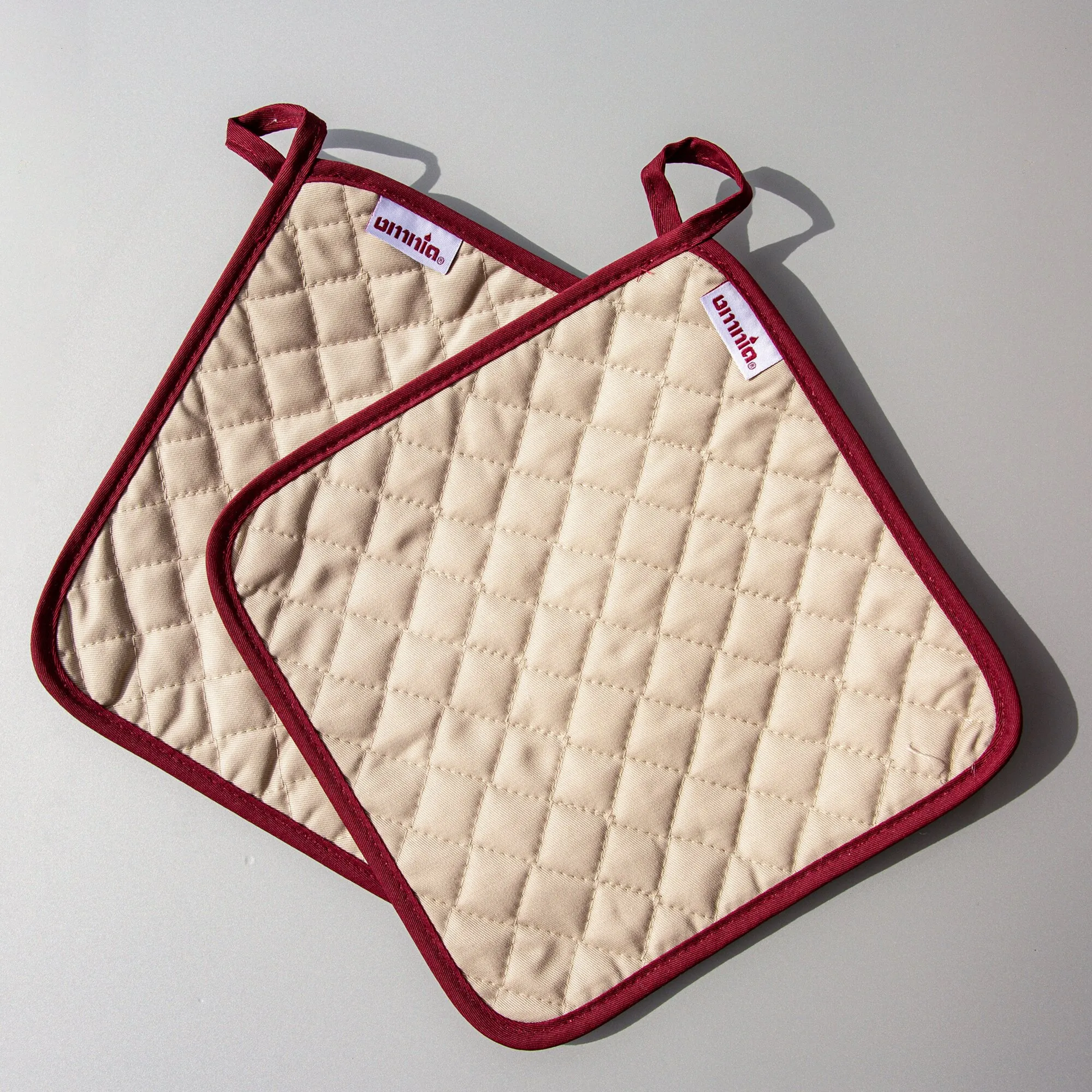 Zwei beige Topflappen mit karierter Steppung, bordeauxroter Einfassung und Aufhängeschlaufen auf hellem Untergrund