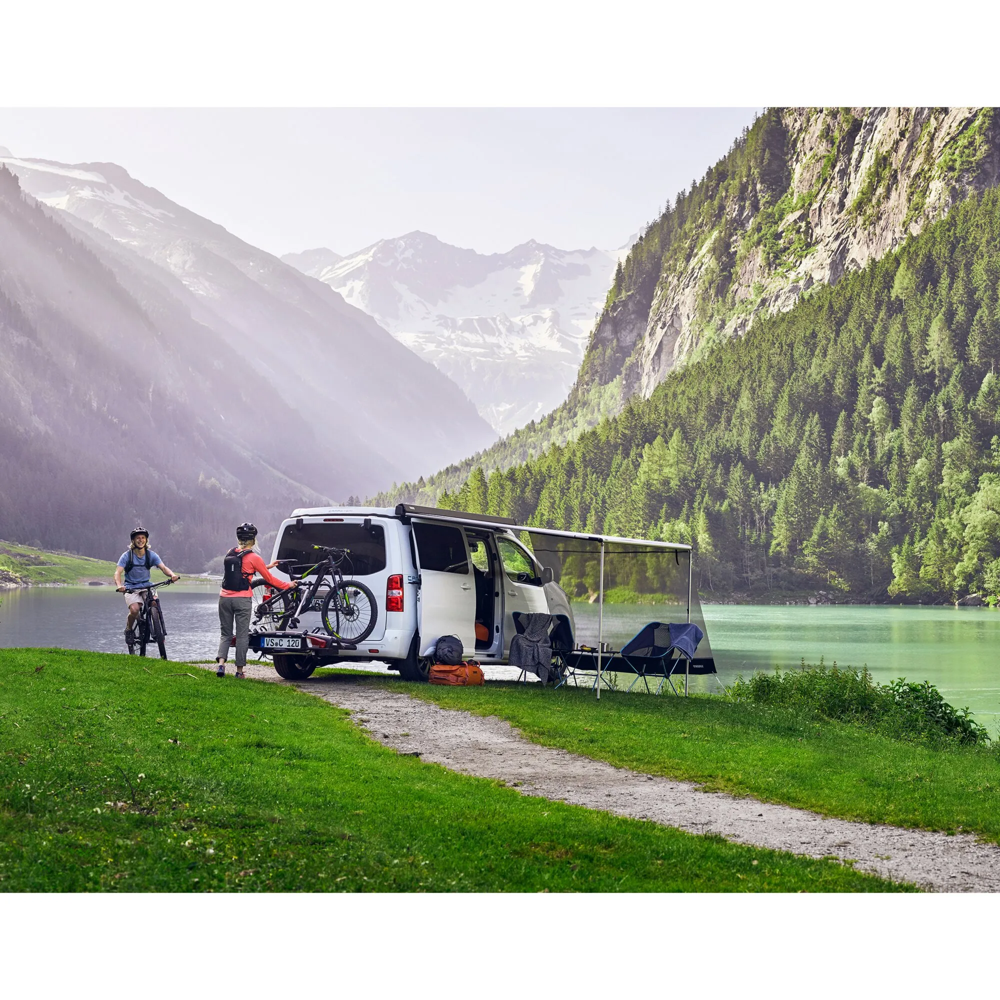 Weiße Campervan mit ausgefahrener anthrazitfarbener Thule-3200-Markise am Bergsee, Fahrrädern und zwei Radfahrern
