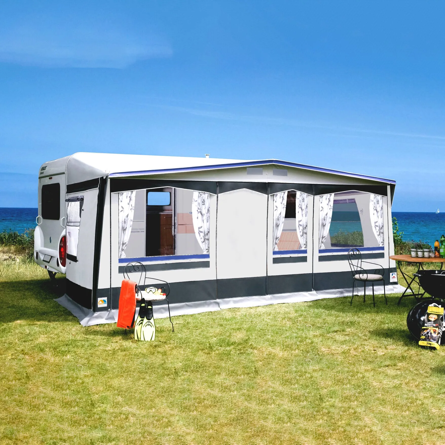 Weiß-graues Saisonvorzelt Prestige mit großen Fenstern und Vorhängen, an einem Wohnwagen am Meer aufgebaut, mit Sitzmöbeln davor